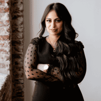 A confident woman with long hair wearing a black dress with sheer polka-dot sleeves standing with her arms crossed in an indoor setting with a brick wall backdrop.