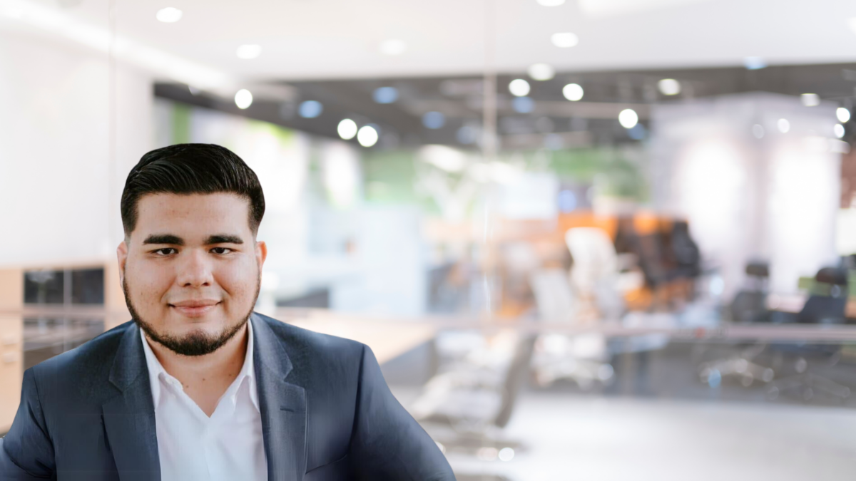 A man with dark hair and a beard, wearing a navy blue suit and white shirt, is smiling and looking at the camera. He is sitting in an office environment with blurred background elements including desks, chairs, and office equipment.