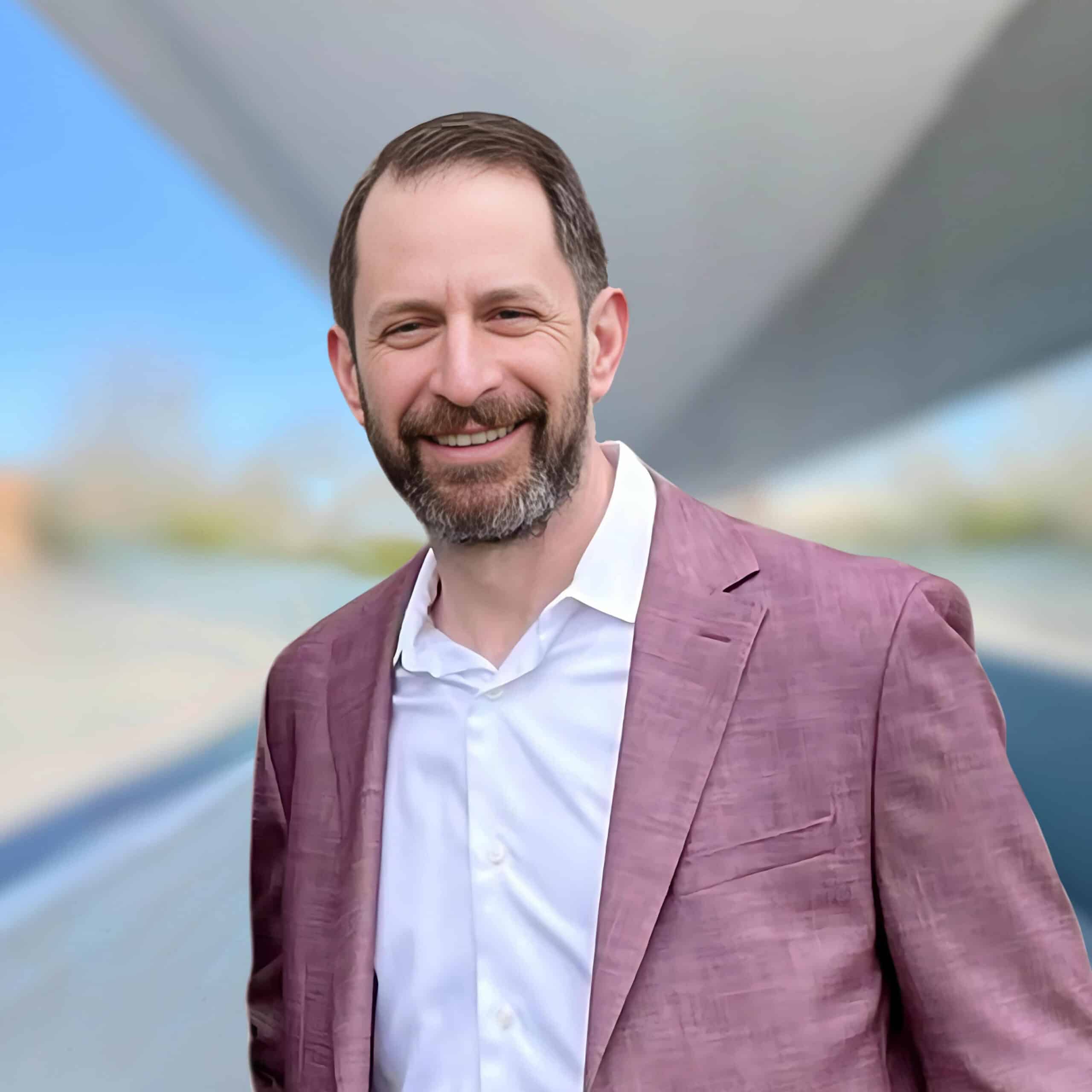 A man with short brown hair and a beard is smiling at the camera. He is wearing a white shirt beneath a mauve blazer. The background is abstract with blurred patterns and shades of blue and grey.