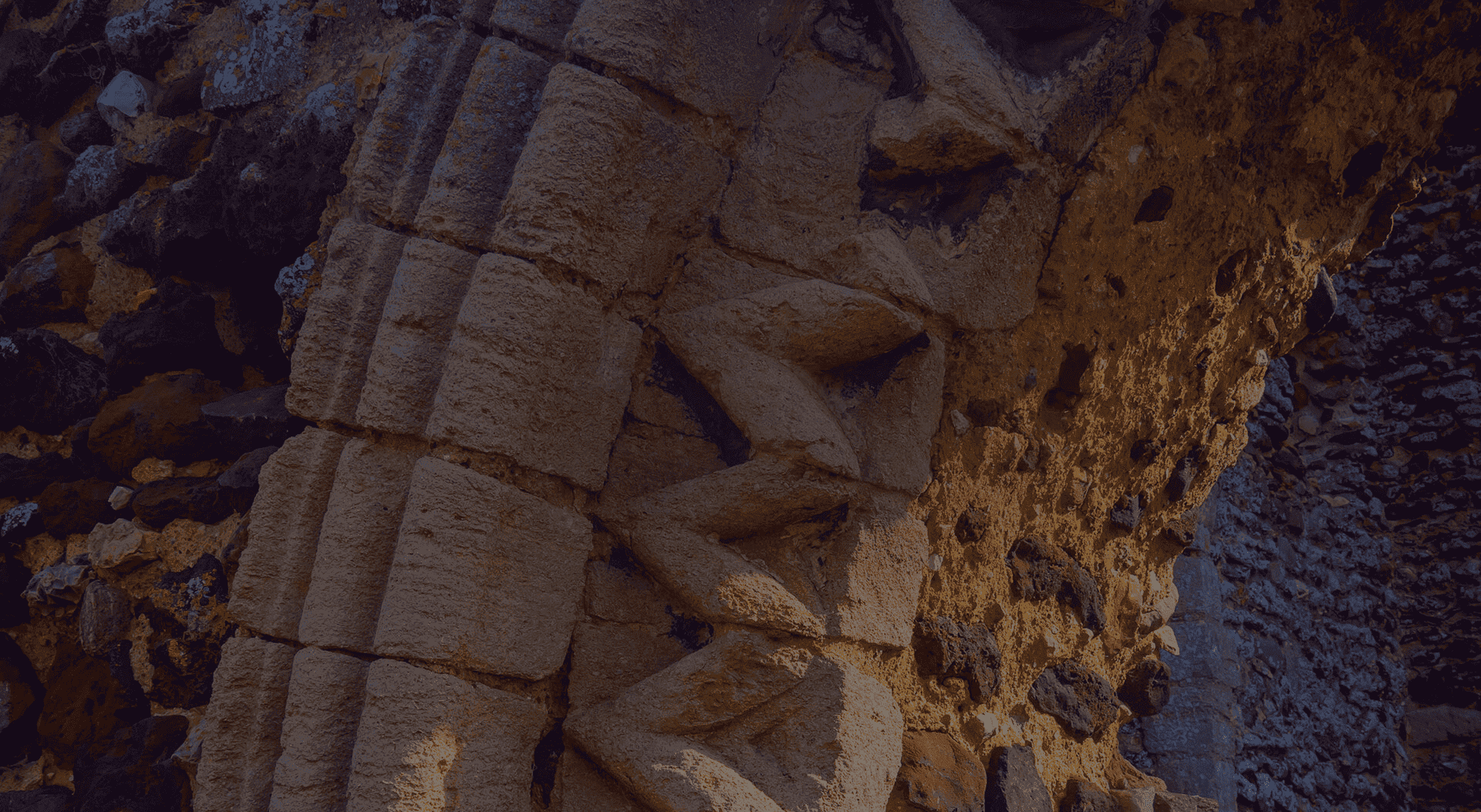 Close-up of an ancient stone archway, featuring intricately carved geometric patterns. The stone has a weathered appearance with a mix of light and dark shades, illuminated by a warm light, suggesting sunset or artificial lighting.