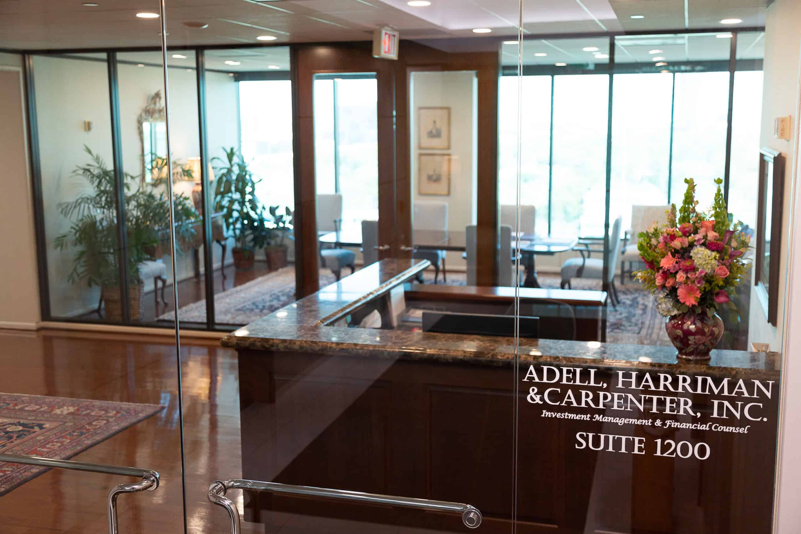 A view through glass doors into a professional office suite with a reception desk, a floral arrangement, seating, plants, and large windows. The door is labeled “Adell, Harriman & Carpenter, Inc., Suite 1200.”.