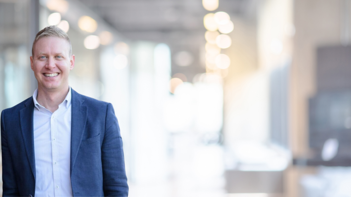 A smiling man in a blue blazer and white shirt stands indoors with a modern, brightly lit office corridor blurred in the background.