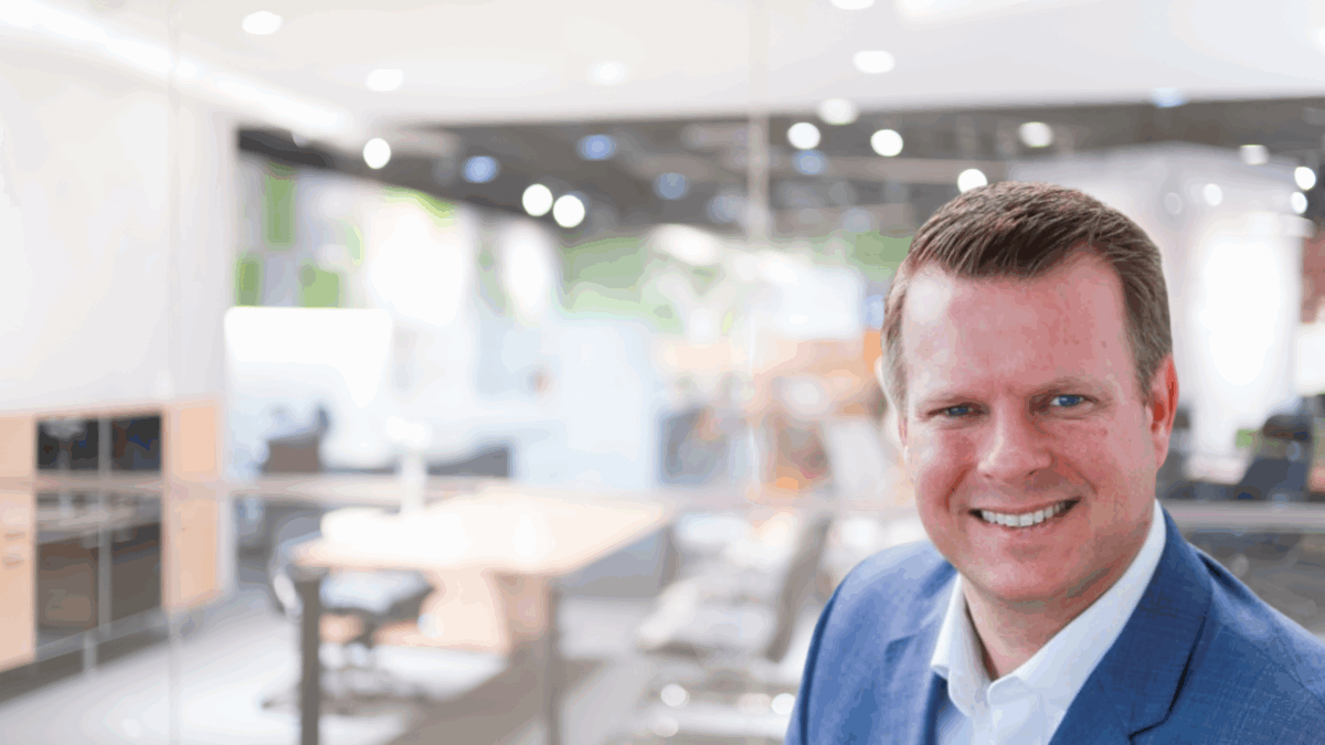 A smiling man in a blue suit stands in the foreground of a modern, brightly lit office with glass walls and blurred desks in the background.