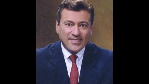 A man wearing a dark suit, white shirt, and red tie is smiling at the camera against a plain brown background.