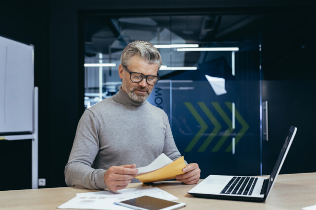 A man with gray hair and glasses, wearing a gray turtleneck, sits at a desk reviewing papers. A laptop, tablet, and documents are on the desk in a modern office with glass walls and a whiteboard.