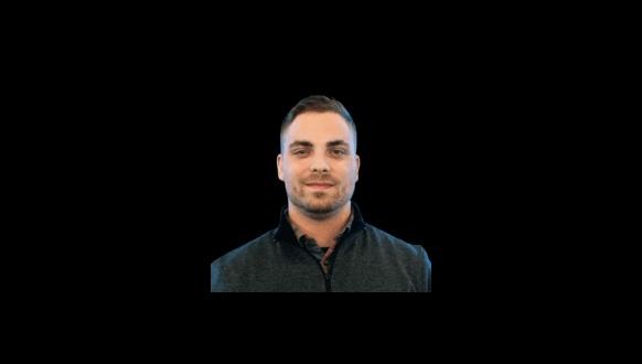 A man with short dark hair and light skin is facing forward, smiling slightly. He is wearing a dark-colored zip-up jacket. The background is solid black.