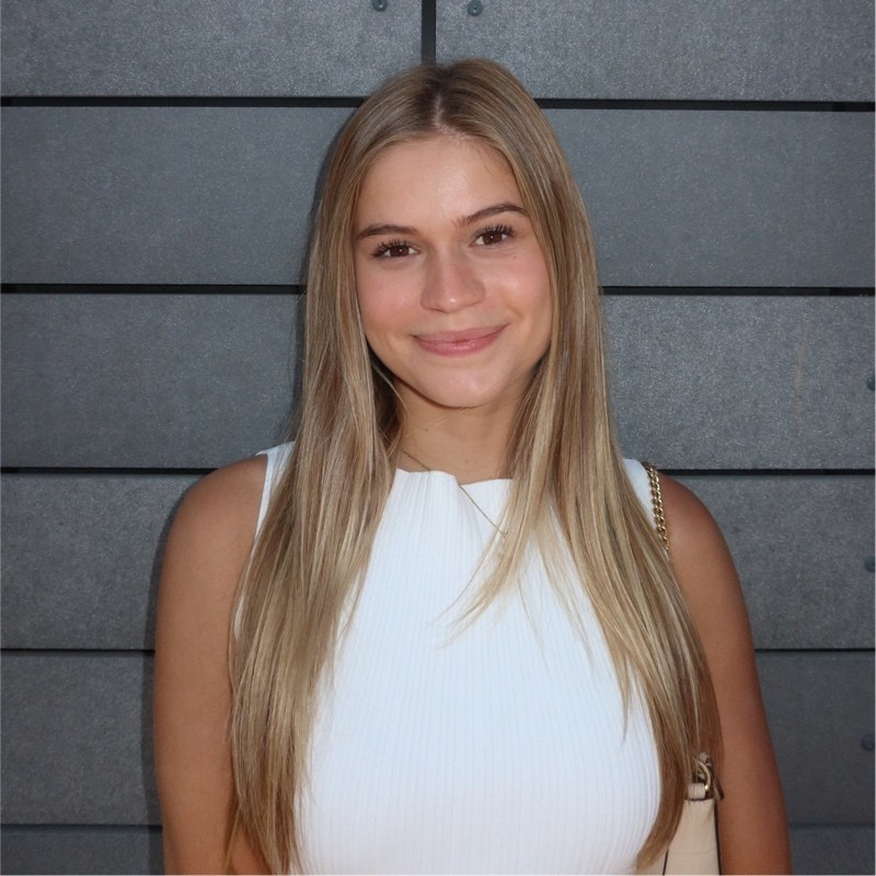 A young woman, Jacquelyn Elliott, with long, straight blonde hair and a white sleeveless top smiles at the camera while standing in front of a gray, horizontally paneled wall.