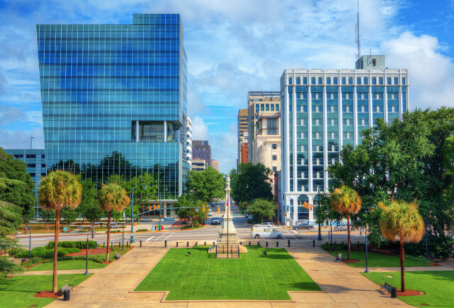 A cityscape featuring a central green plaza with palm trees, a tall obelisk monument, and modern glass and historic stone buildings under a blue, partly cloudy sky.