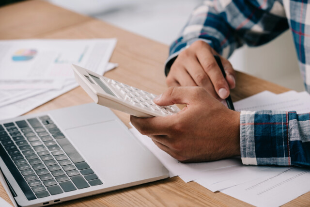 A person in a plaid shirt uses a calculator at a wooden desk with a laptop, pen, and paperwork scattered around, suggesting work or financial calculations.