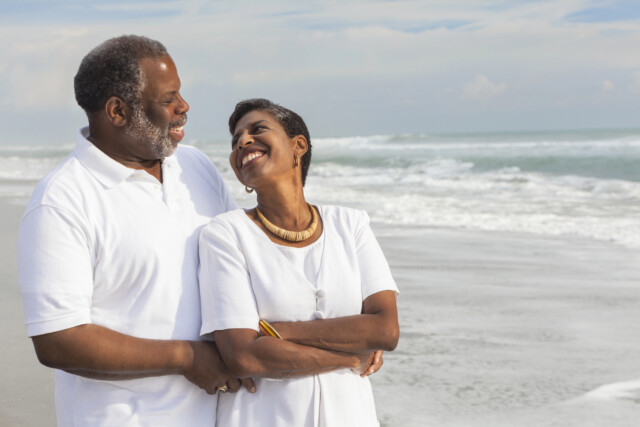 An older couple smiles and embraces on a beach, both wearing white clothes. The ocean and waves are in the background under a partly cloudy sky. They appear happy and relaxed.