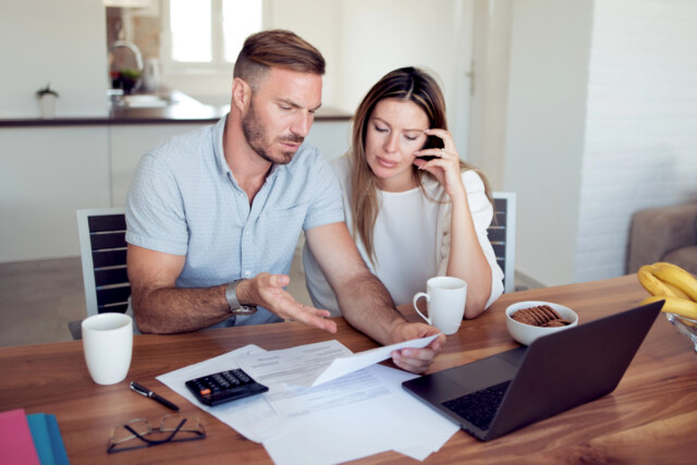 A couple sits at a table with paperwork, a laptop, calculator, and coffee mugs. The man gestures while looking at documents, and the woman is talking on the phone, appearing concerned.