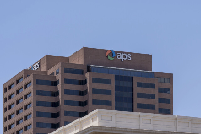 A brown high-rise office building with many windows displays the "aps" logo and name near the top, set against a clear blue sky. A lighter colored building is partially visible in the foreground.