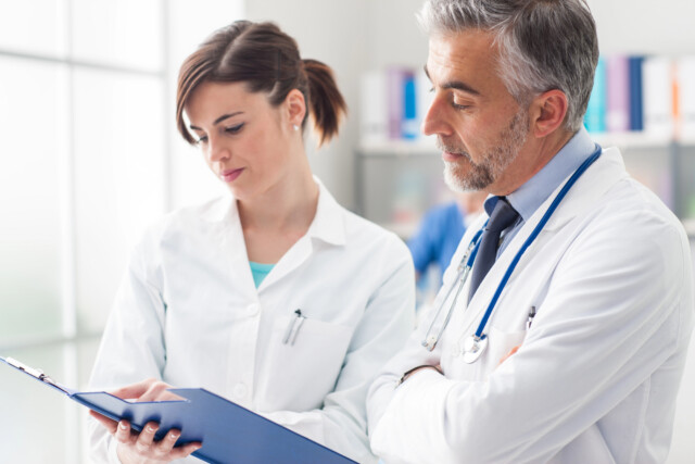Two doctors in white coats are standing together in a bright office, reviewing a document on a blue clipboard and discussing medical information. One has a stethoscope around his neck; shelves are visible in the background.