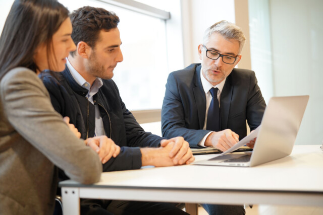 Three people sit at a table in an office, looking at a laptop. An older man in a suit gestures while speaking to a younger man and woman, who are attentively listening and sitting side by side.