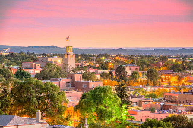 A colorful sunset sky glows over a cityscape with adobe-style buildings, lush green trees, and a prominent clock tower with a flag, set against distant hills. City lights begin to illuminate the scene.