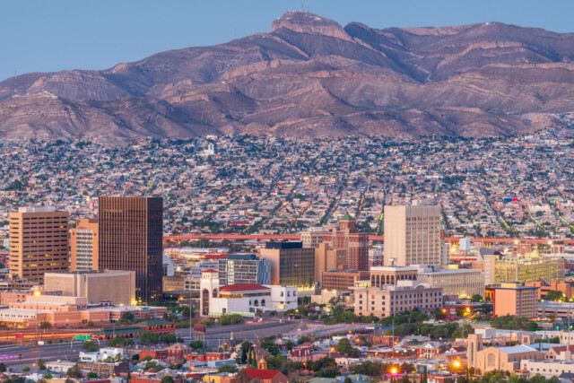 Downtown cityscape with tall buildings in front of a sprawling residential area, set against large, rugged mountains at dusk under a clear sky.