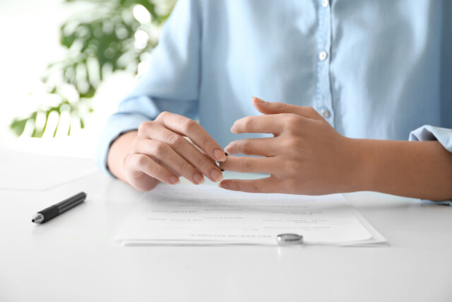 A person in a blue shirt removes a ring while sitting at a desk with divorce papers and a pen, suggesting the end of a marriage.